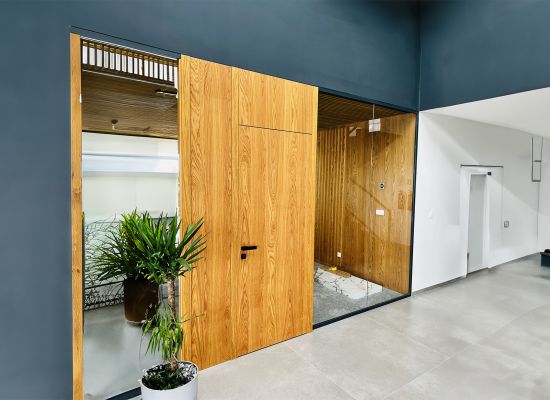 Kliknutím zvětšíte Dřevěné dveře SAPELI s dřevěným obložením ve stejném dekoru vsazené do skleněné stěny. Elegantní kombinace skla a dřeva podtrhuje minimalistický design prostoru a je chloubou nových kanceláří.